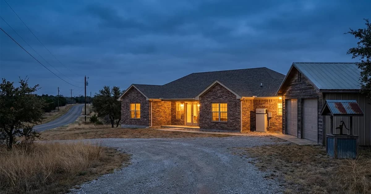 A single-story brick ranch home on open acreage west of Houston at blue hour, warm light in the windows, a wall-mounted home battery and a small well pump house visible beside a detached garage, with a long rural road and distant utility poles behind it.